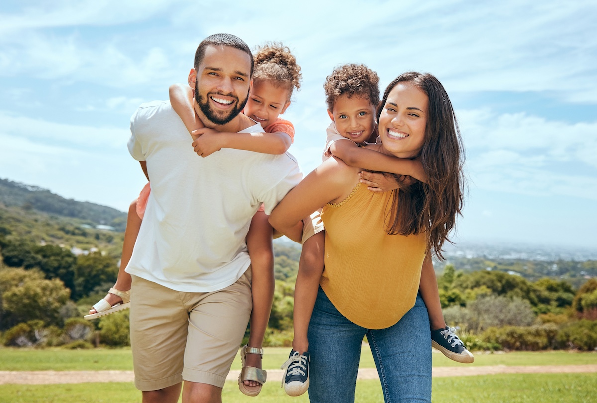 Active kids, happy family. Mom, dad and children on piggyback ride from parents in nature park for fun, summer time bonding and outdoor family activity. Black father, mother and kids smile together while playing on grass.