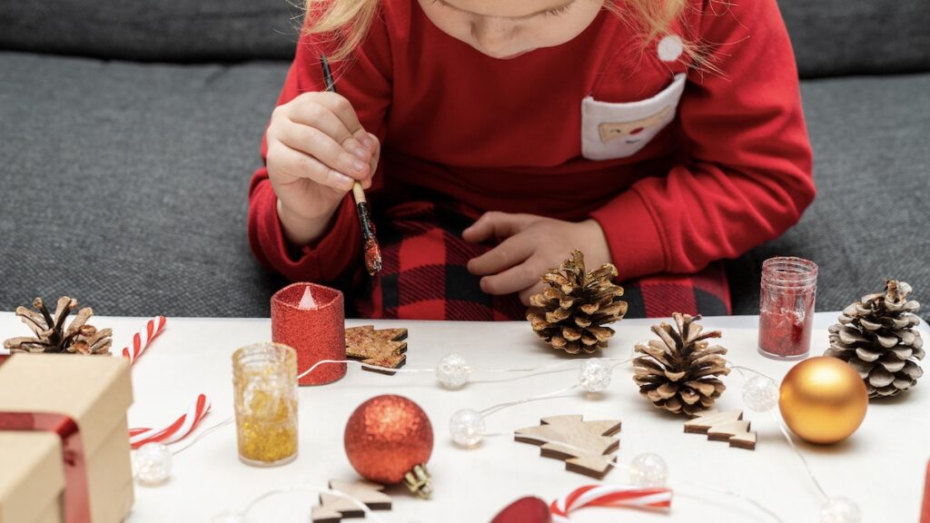 Little girl painting pine cones and wooden Christmas tree decorations at home