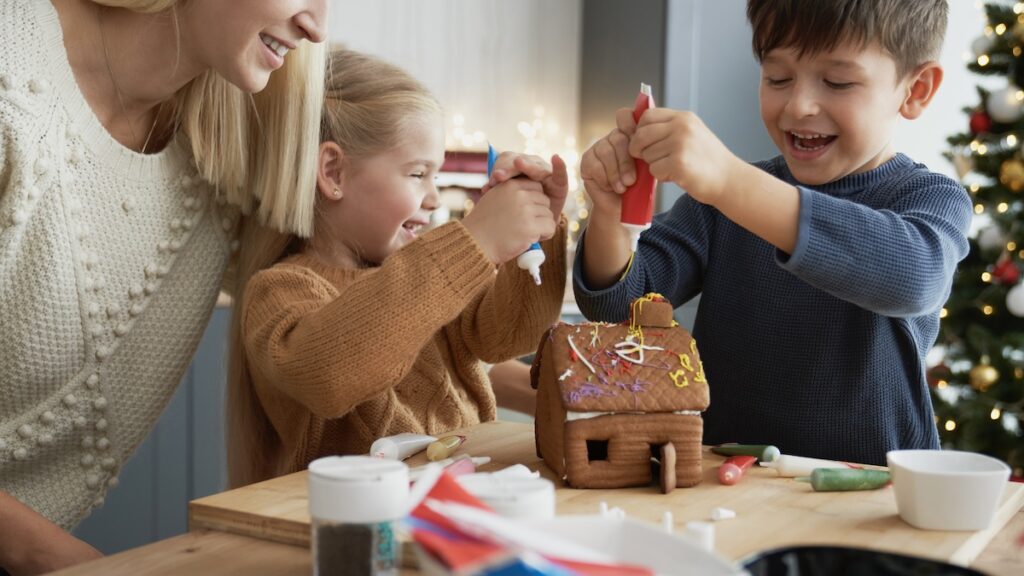 One of 10 Fun Christmas Activities for Kids to Make the Season Magical - Mom and two little children having fun while decorating a gingerbread house