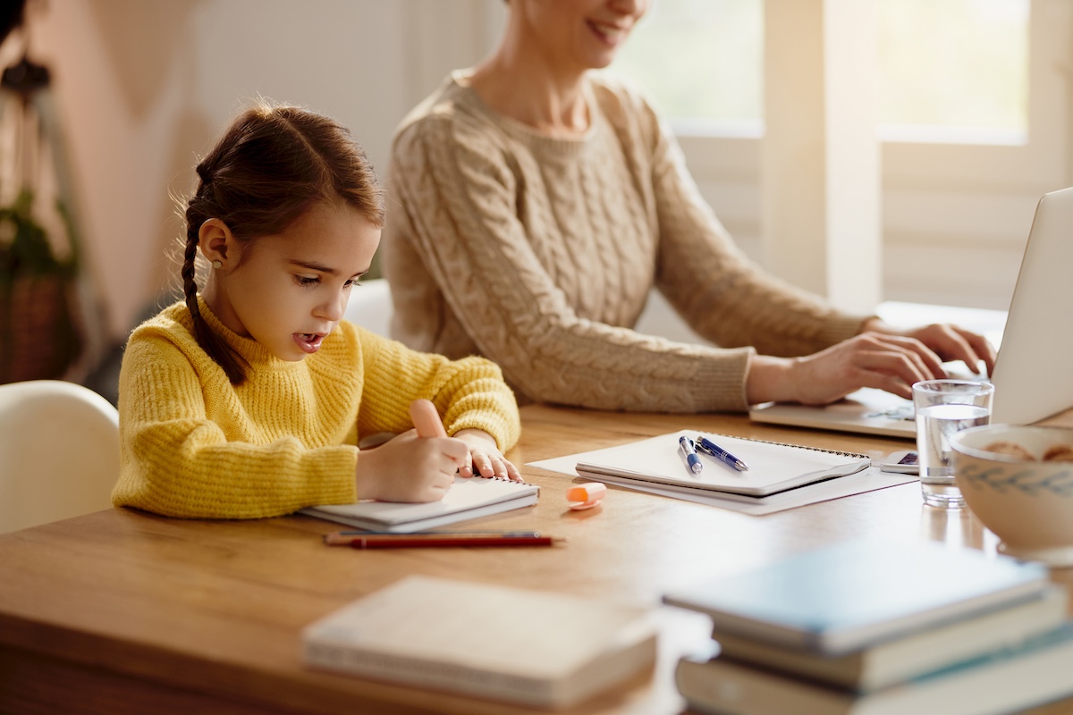 Little girl doing homework while mother is working on a computer at home.