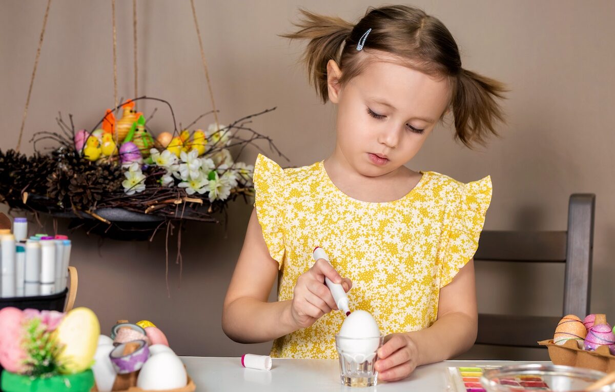 Fun and Creative Easter Activities for Kids, Little adorable girl in yellow dress painting Easter eggs with paintbrush